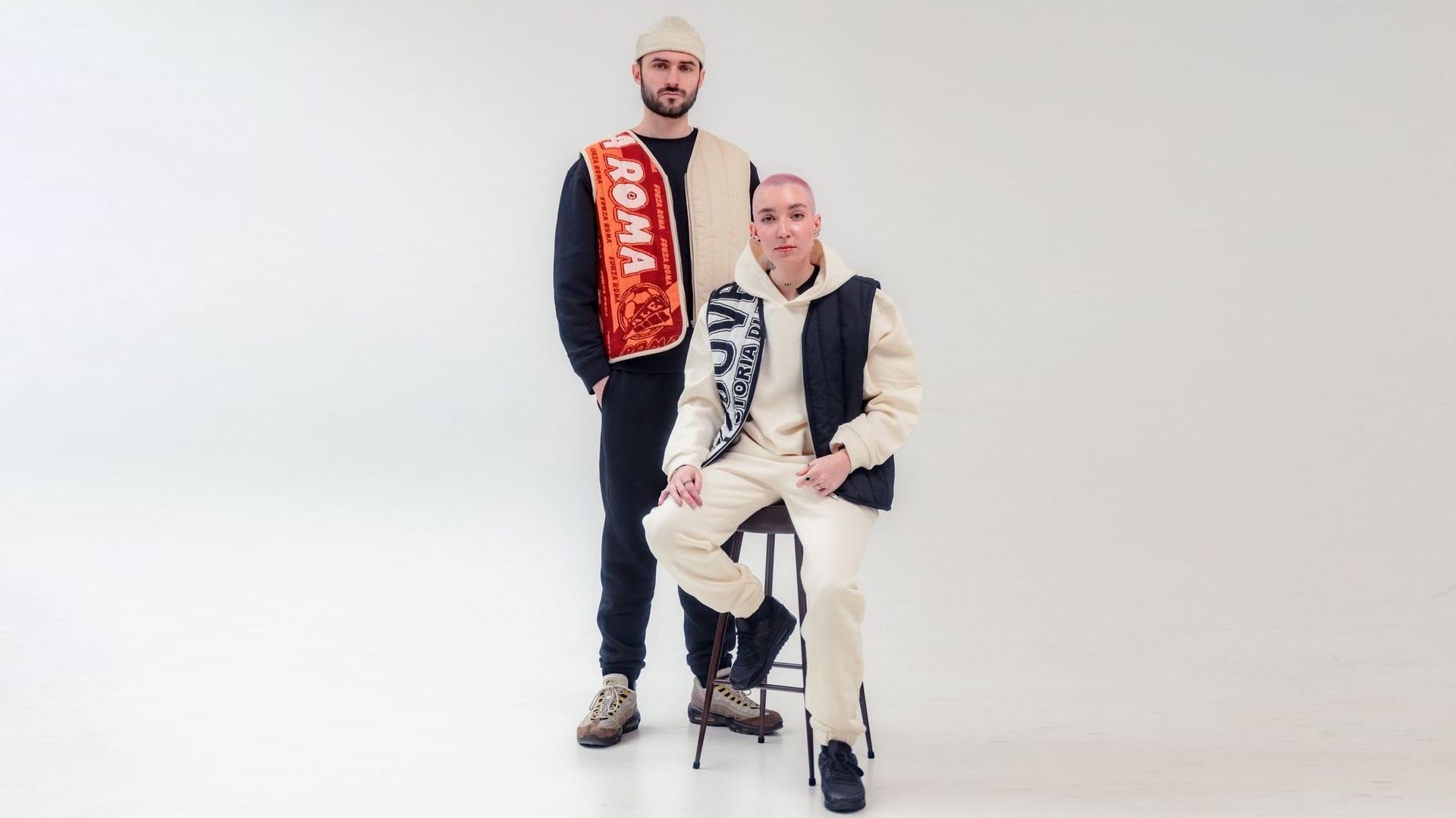 Two models in a white studio space wearing football-inspired streetwear with scarves draped over their shoulders. One is seated in a cream tracksuit and vest, the other stands in black and beige with an orange “ROMA” scarf.